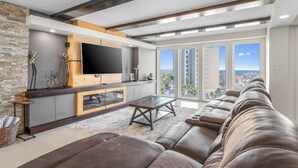 A modern living room with a large leather sofa, a flat-screen TV mounted on a wooden panel, and a large window offering a city view. - Amazing Panoramic Ocean Paradise (South Padre Island)