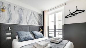A modern bedroom with a marble-patterned headboard, a large window with curtains, a black hanger on the wall, and neatly folded towels on the bed.