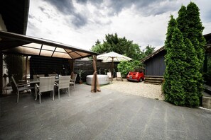 Un espace extérieur aménagé en terrasse, avec un coin salon couvert, une voiture rouge d’époque et un grand arbre à feuilles persistantes.