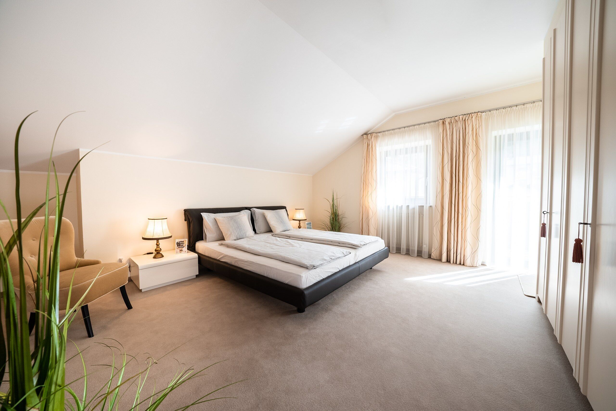 Un dormitorio con cama, mesitas de noche, lámpara, sillón y una planta.