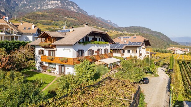 A hillside property with multiple buildings, including a main house with a sloped roof and solar panels, surrounded by vineyards and mountains in the background. - 723A Apartment Burgunderhof (Termeno sulla strada del vino)