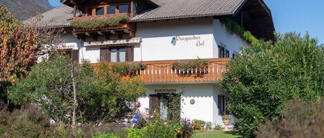 A traditional chalet with a wooden balcony, a mountain backdrop, and a sign indicating it's a Baugut.