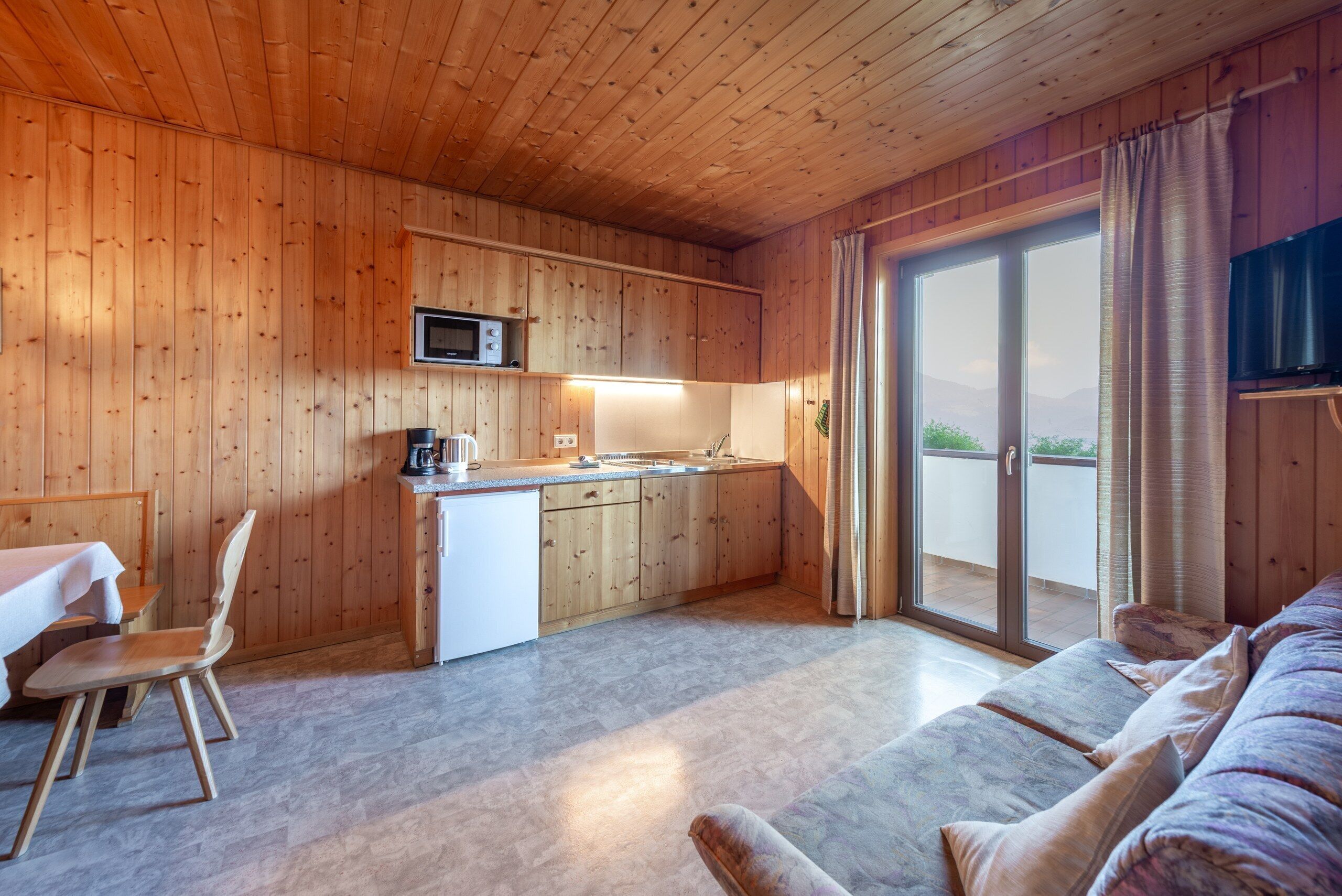 Un intérieur de chalet chaleureux avec des murs en bois, un coin cuisine, un espace repas avec une table et des chaises, et une porte-fenêtre coulissante donnant sur un balcon.
