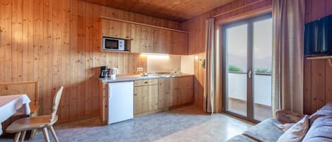 Interior acogedor de una cabaña con paredes de madera, una kitchenette, un comedor con mesa y sillas, y un ventanal corredizo que da a un balcón.