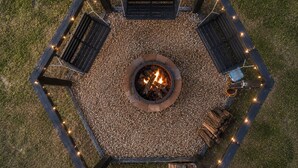 Un espacio de descanso hexagonal al aire libre con un brasero central, rodeado de bancos y luces colgantes.