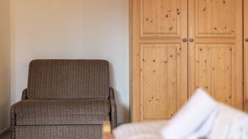A room with a brown upholstered chair, a wooden wardrobe, and a bed with a white pillow.