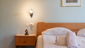 Una cama bien hecha con sábanas blancas, una mesita de noche de madera con una pequeña figurilla, una lámpara de pared y un cuadro en la pared.