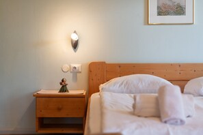 A neatly made bed with white linens, a wooden nightstand with a small figurine, a wall-mounted light, and a framed picture on the wall.