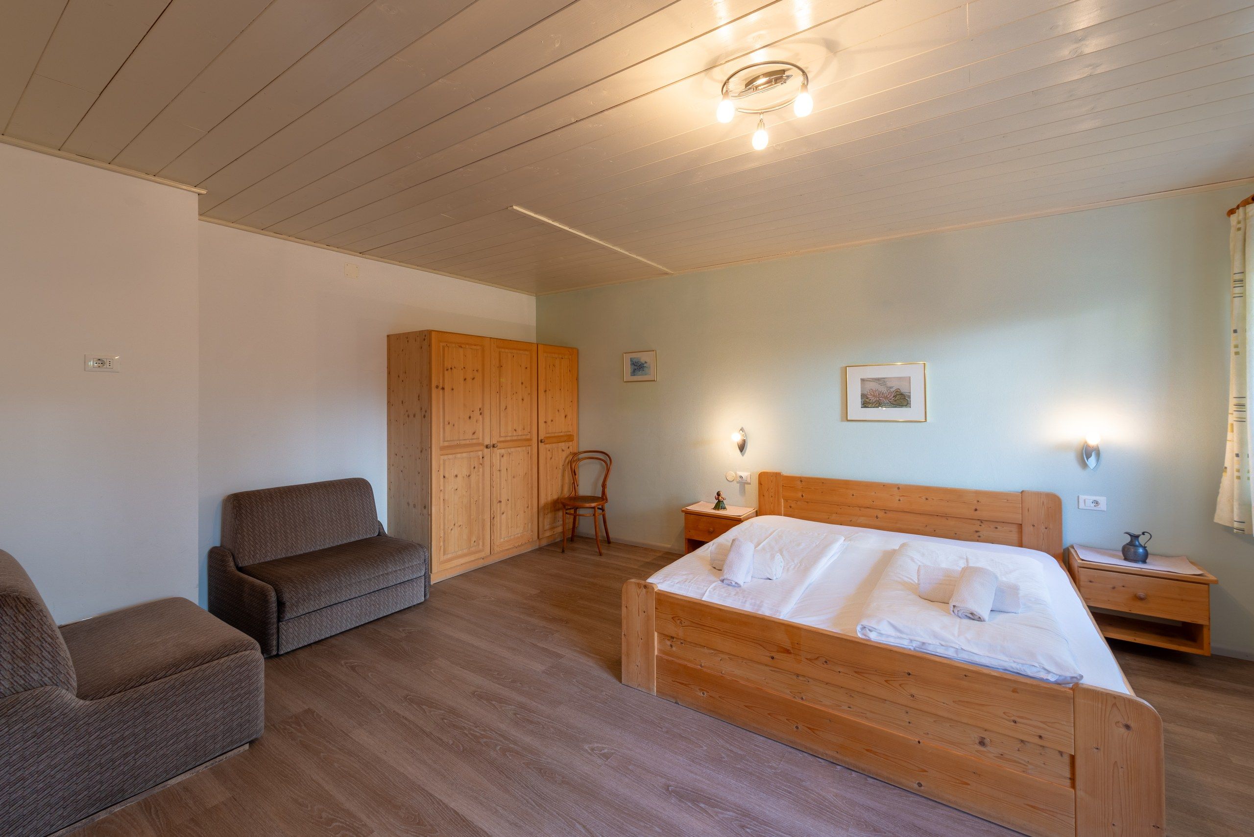 Une chambre à coucher avec un lit en bois, une armoire en bois, une chaise en bois, une table de chevet et un luminaire fixé au mur.