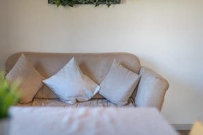 Un sofá beige con tres almohadones frente a una pared lisa, una planta pequeña en primer plano y una mesa blanca con mantel blanco.