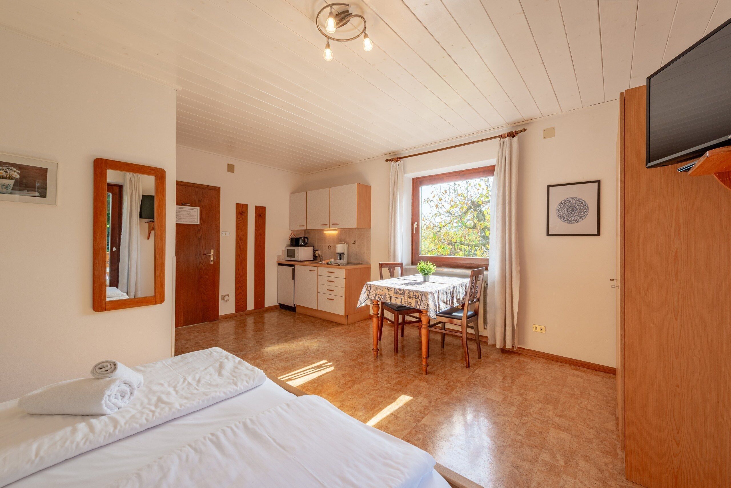 A room with a bed, a dining table with chairs, a kitchen area, and a television mounted on the wall.