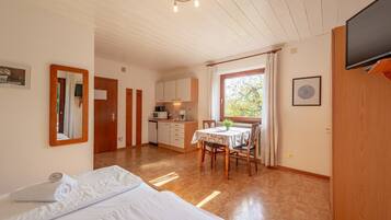 A room with a bed, a dining table with chairs, a kitchen area, and a television mounted on the wall.