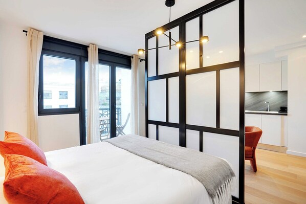 A modern hotel room with a large bed, a balcony with a view, a kitchenette, and a sliding door separating the room from the balcony.