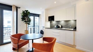 Une cuisine moderne avec des armoires blanches, un comptoir noir et un four à micro-ondes encastré. On y trouve une table ronde avec deux chaises orange et une plante en pot.