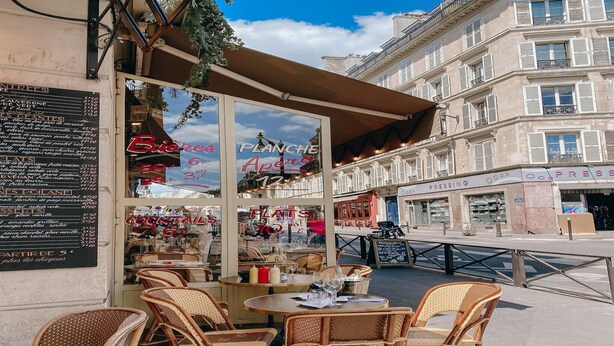 Un café en plein air avec des chaises et des tables en osier, un tableau affichant le menu et une vue sur la rue.