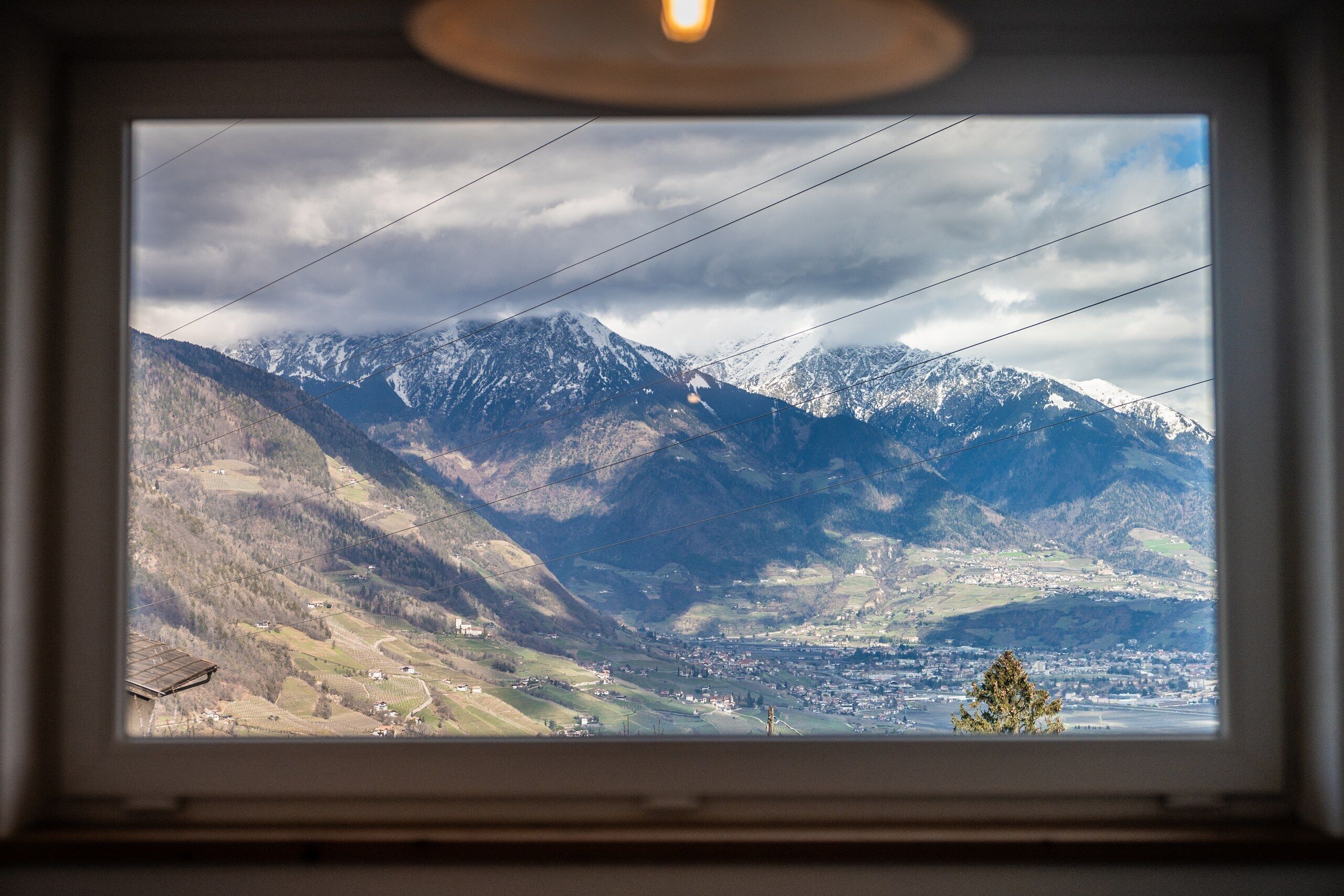 Vista di un paesaggio montuoso attraverso una finestra, con una valle punteggiata di edifici e una vetta montuosa imponente.