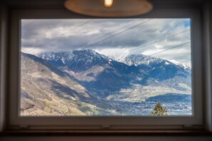 Vista di un paesaggio montuoso attraverso una finestra, con una valle punteggiata di edifici e una vetta montuosa imponente.