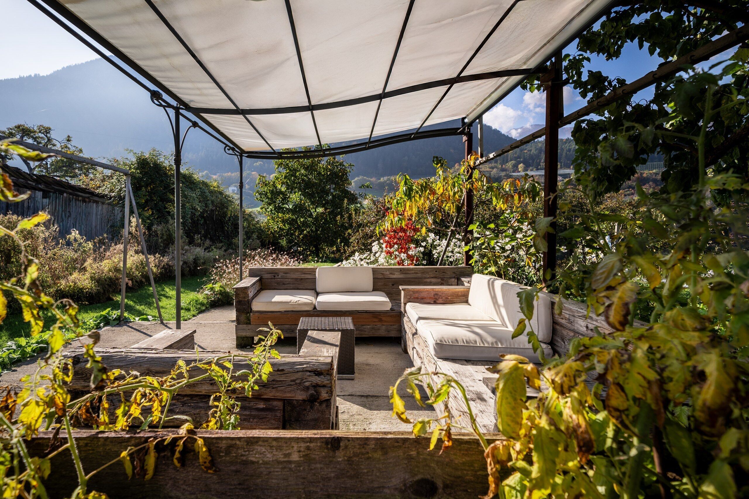 Ein überdachter Außenbereich mit einer Couch und Stühlen, umgeben von Pflanzen und Blick auf eine gebirgige Landschaft.