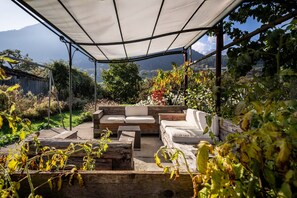Ein überdachter Außenbereich mit einer Couch und Stühlen, umgeben von Pflanzen und Blick auf eine gebirgige Landschaft.