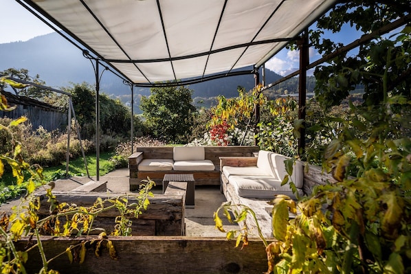 A covered outdoor seating area with a sofa and chairs, surrounded by plants and a view of a mountainous landscape.