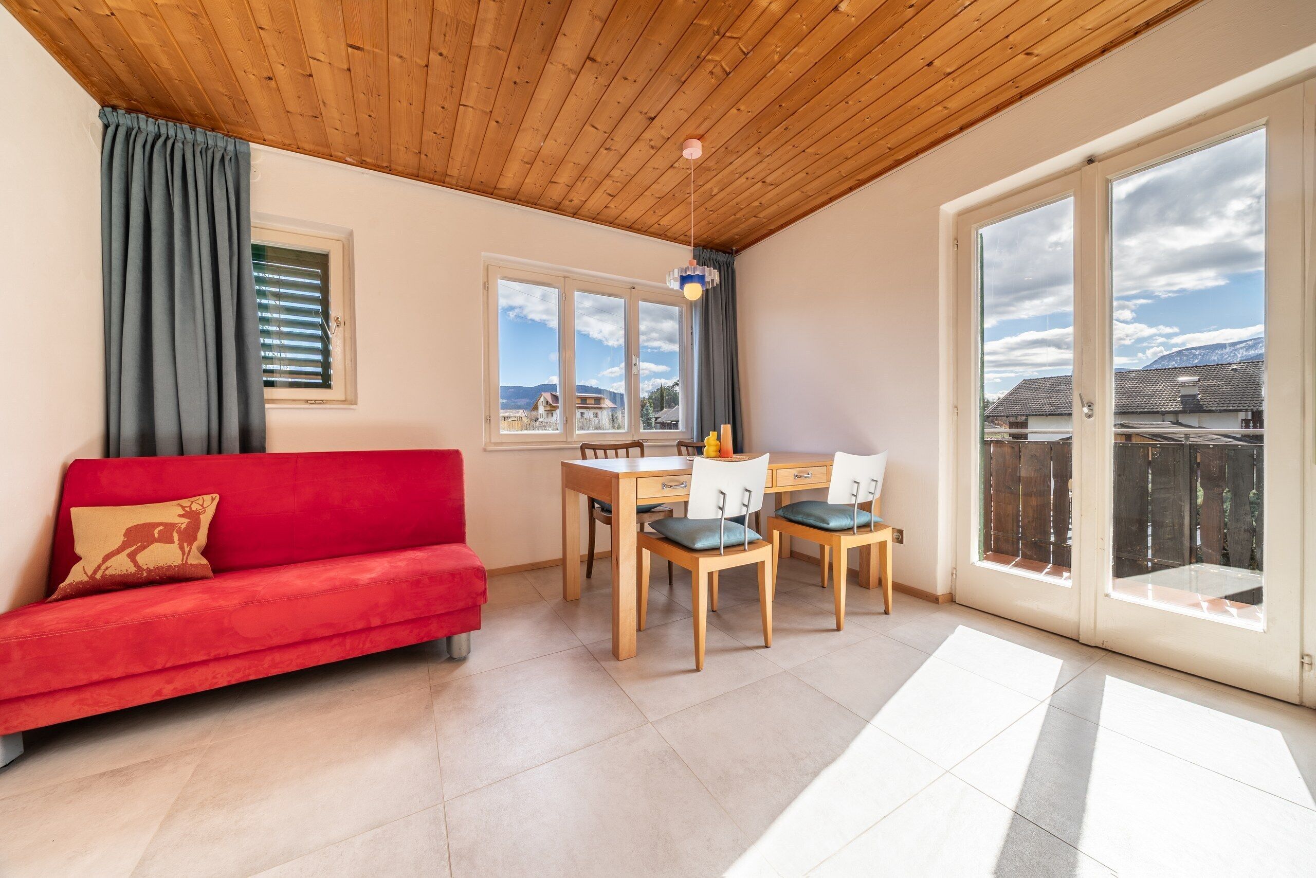 A cozy room with a red sofa, a wooden dining table with chairs, and large windows offering a view of a mountain landscape.