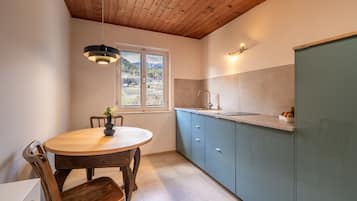 Una cucina con soffitto in legno, un tavolo da pranzo rotondo con sedie, una finestra vista montagne e un lavello con rubinetto.