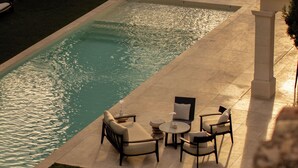 Un área junto a la piscina con sillones de descanso y mesas, una piscina cristalina y un césped bien cuidado.