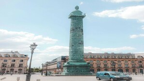 A tall, narrow monument with intricate carvings, surrounded by historic buildings and a clear sky.