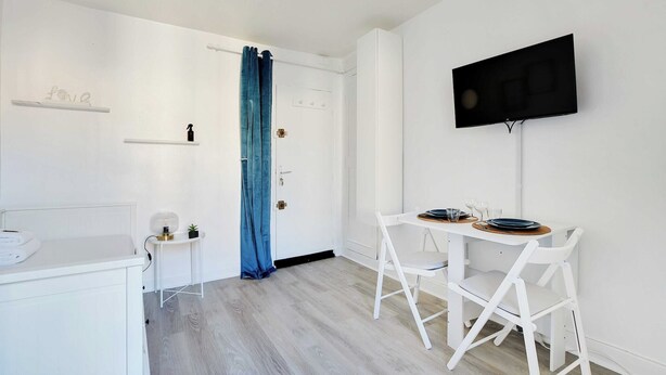 A minimalist room with a white desk, a small table with chairs, a flat-screen TV mounted on the wall, and a blue curtain on the door.