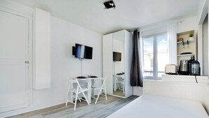 A modern hotel room with a bed, a dining table with chairs, a TV mounted on the wall, a large mirror, and a window with curtains.