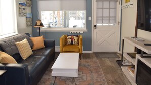 Un salon avec un canapé en cuir noir, un fauteuil jaune, une table basse blanche et un téléviseur posé sur un meuble en bois.
