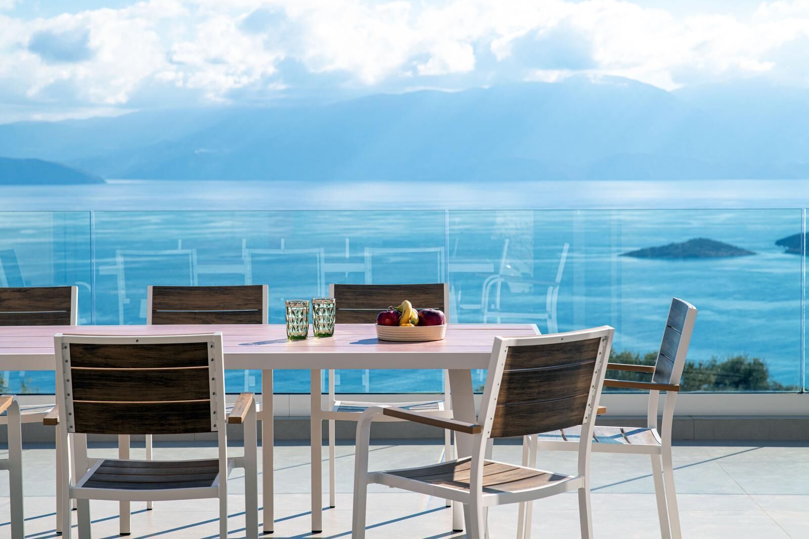 A dining table with chairs, a bowl of fruit, and two glasses on a terrace with a view of the sea and mountains.