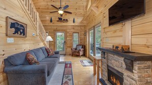 A log cabin interior with a fireplace, a sofa, a ceiling fan, and a welcome sign.