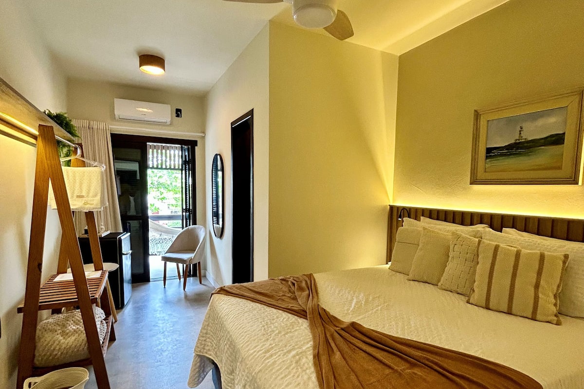 A hotel room with a bed, a ceiling fan, a painting on the wall, and a view of the outdoors.