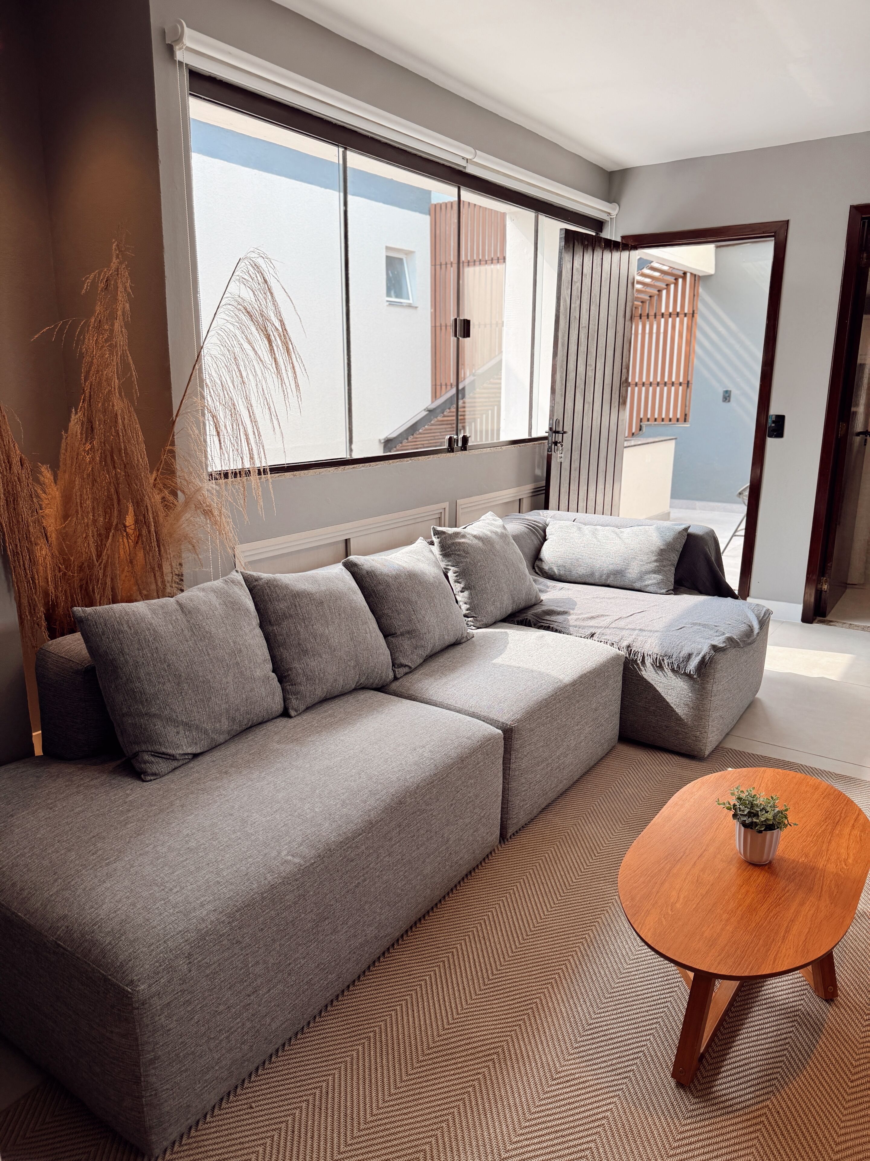 A modern living room with a grey sectional sofa, a wooden coffee table, and a large window with a view of a building.