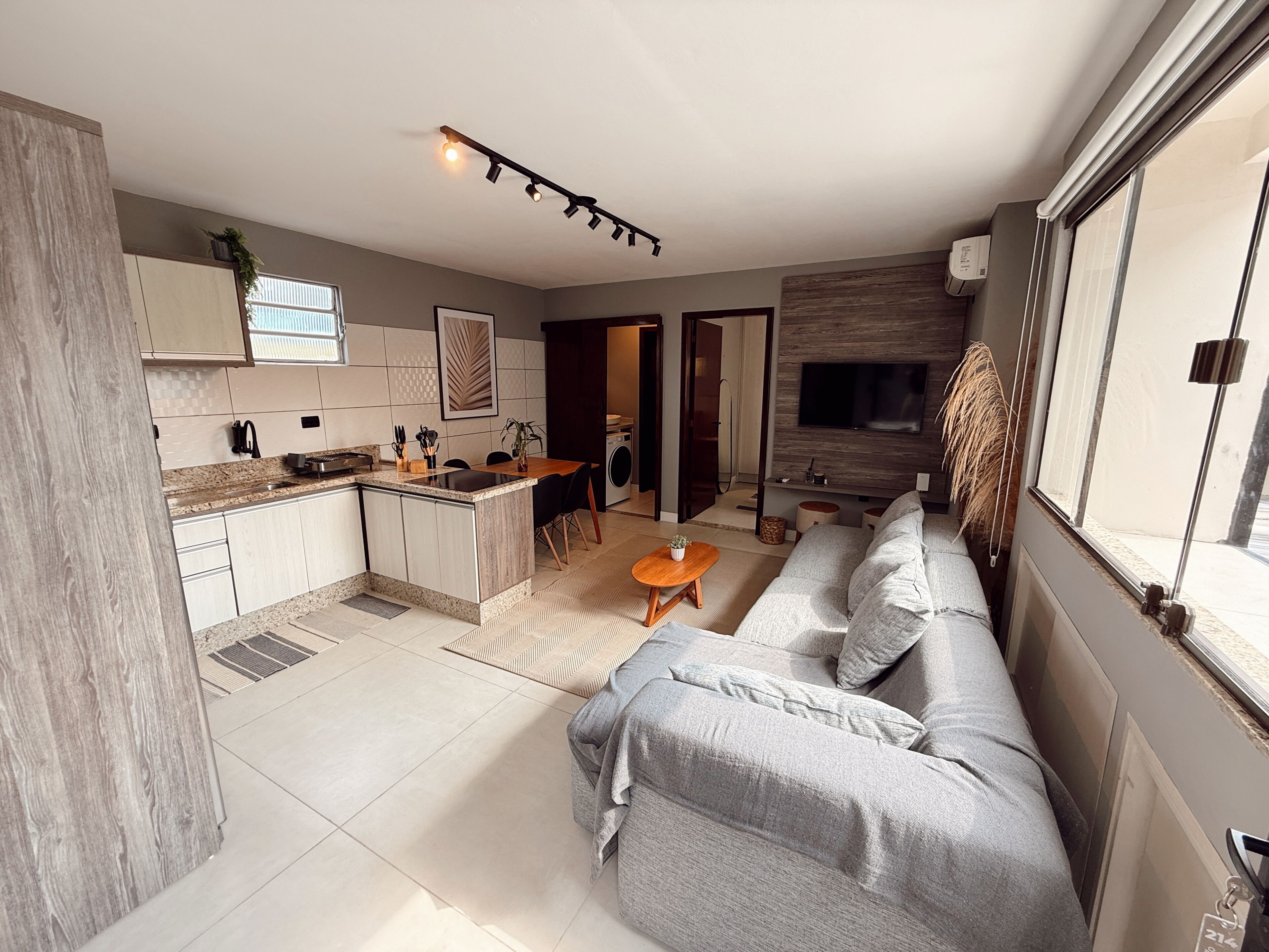 Una cocina moderna con una isla central, una zona de comedor con sofá y mesa, y una sala de estar con televisor y ventanas amplias.
