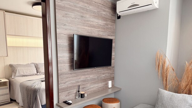 A modern bedroom with a TV mounted on a wooden wall, a bedside table, a bed, and a small seating area with stools. - Pousada Casa Californiana (Garopaba)