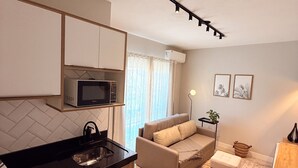 A modern kitchen with a microwave, a sink, and a countertop. There is a beige sofa and a small table with a plant. The room has a tiled floor and a ceiling light fixture. - Pousada Casa Californiana (Garopaba)