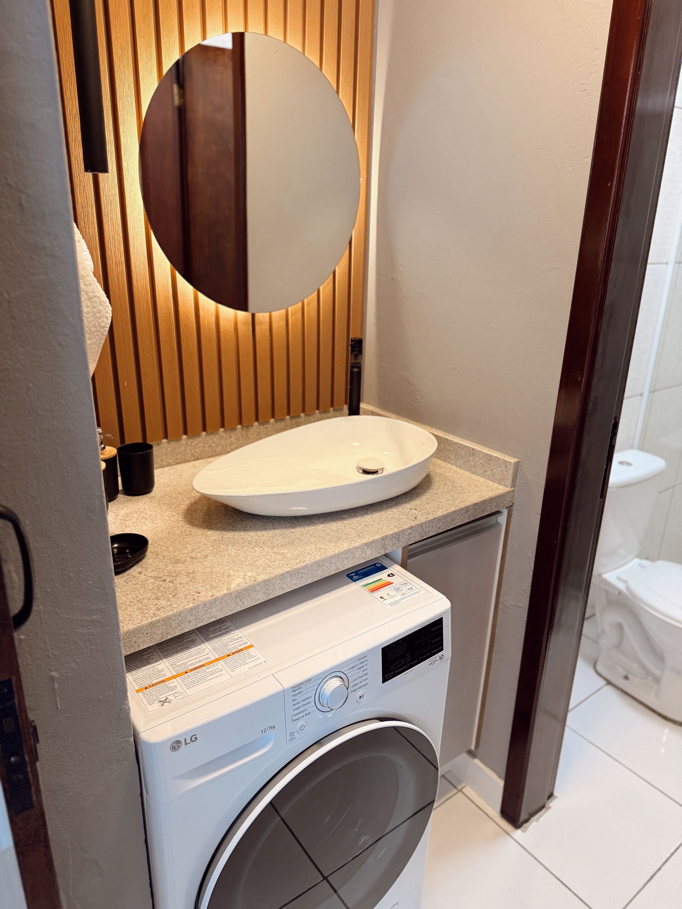 Un baño pequeño con lavadora integrada, un lavamanos con espejo redondo y un inodoro.