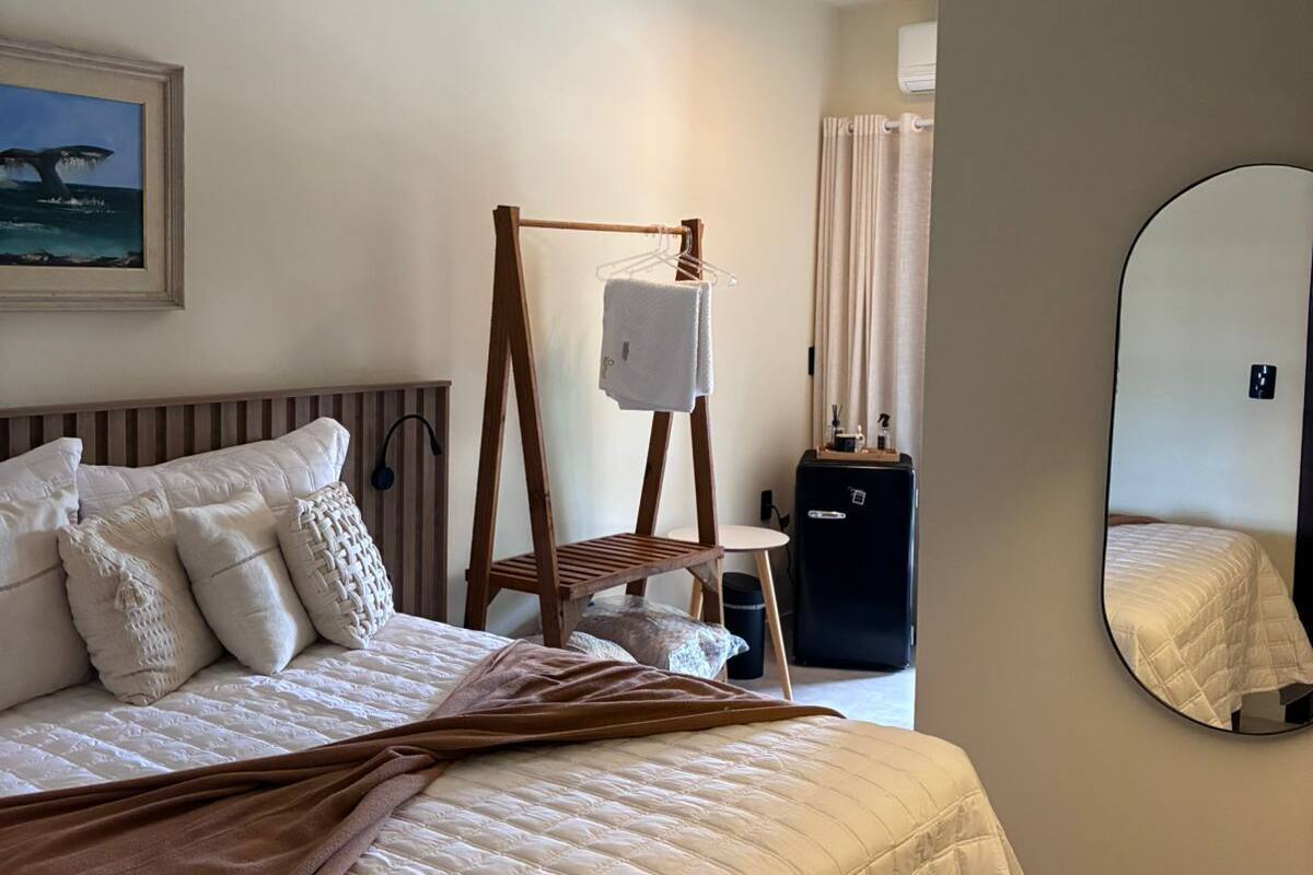 A hotel room with a bed, a ceiling fan, a wooden chair, a small table, a mirror, and a painting on the wall.