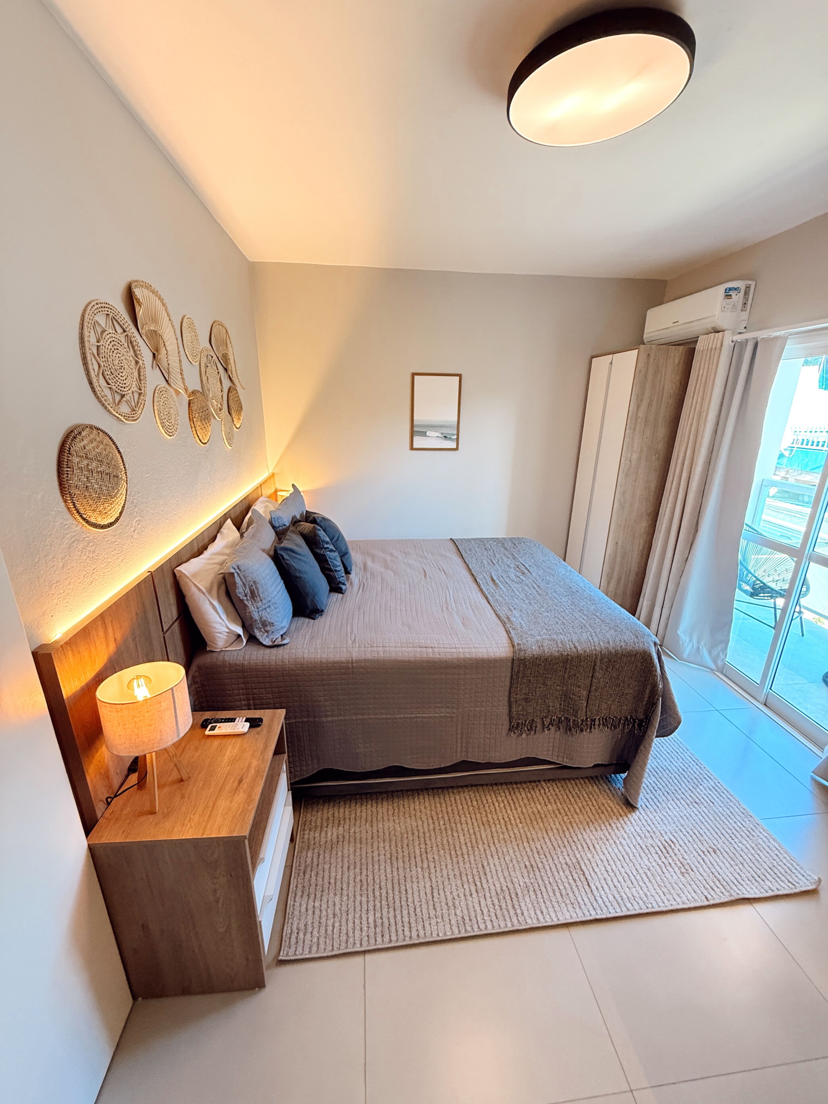 A modern bedroom with a bed, bedside table, lamp, wall art, and a view through the window.
