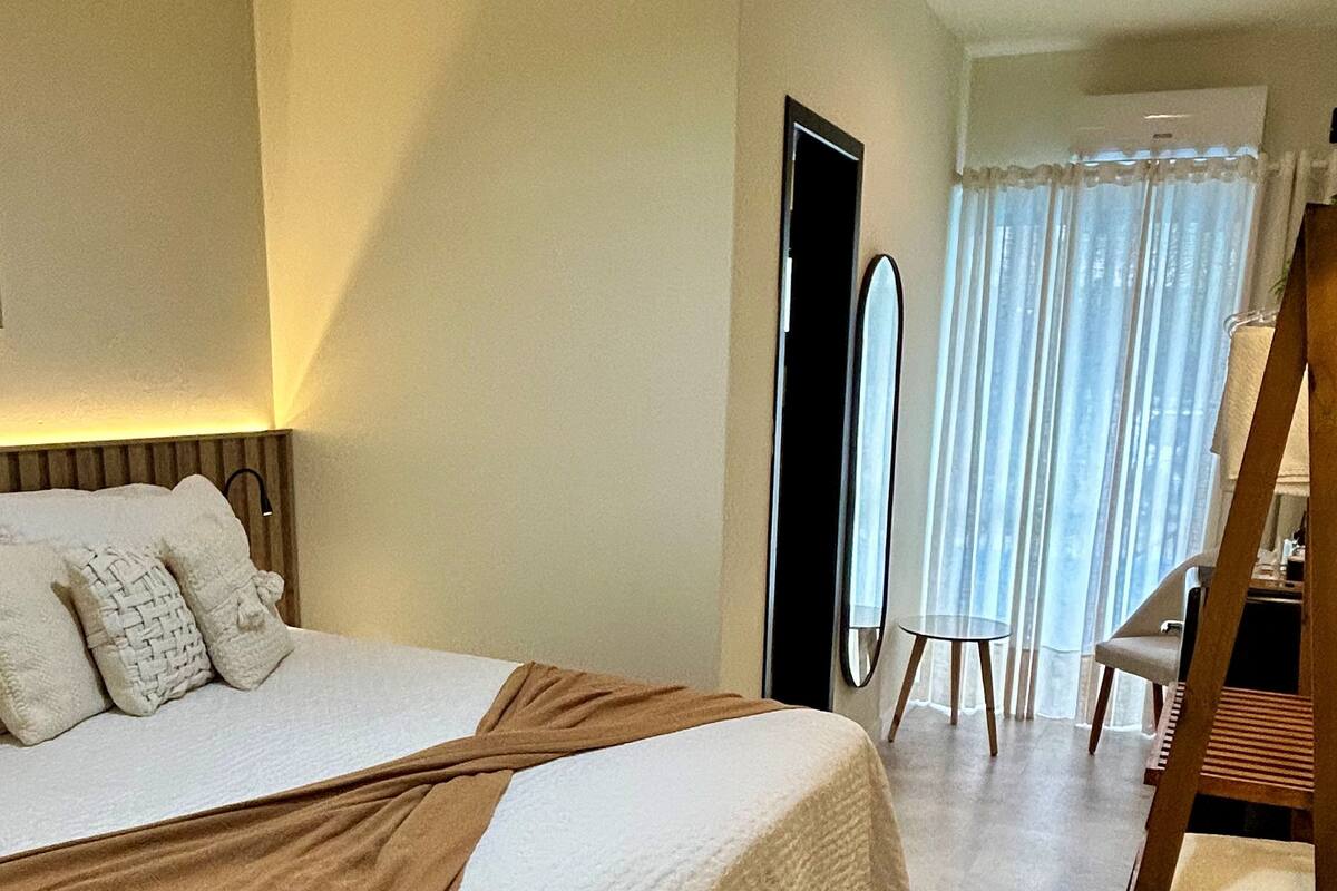 A bedroom with a large bed, a wooden chair, a round woven rug, and a view of a balcony with curtains.
