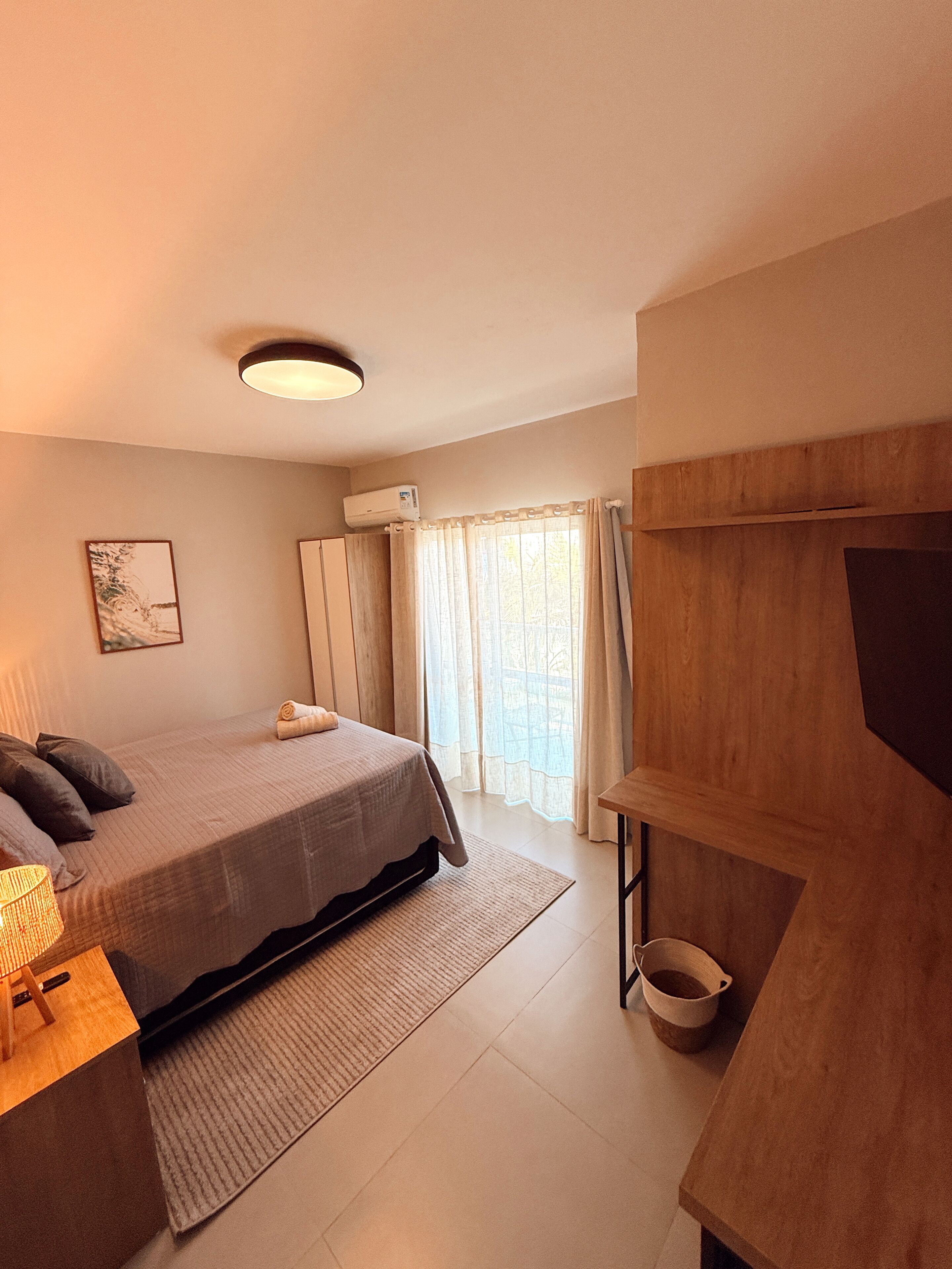 A bedroom with a bed, a bedside table, a lamp, a window with curtains, and a wall-mounted air conditioner.
