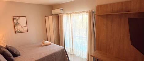 A bedroom with a bed, a bedside table, a lamp, a window with curtains, and a wall-mounted air conditioner.