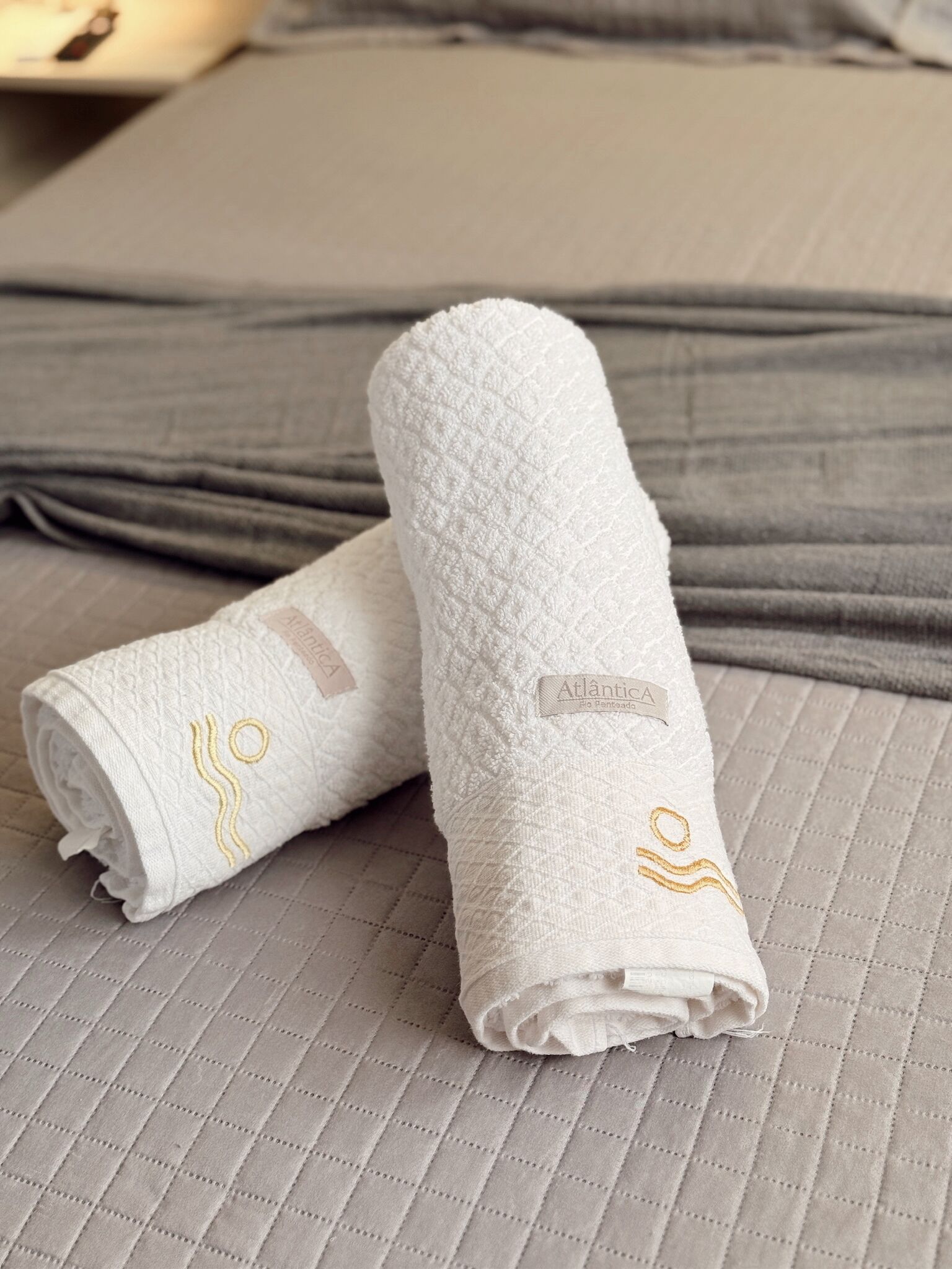 Zwei gerollte weiße Handtücher mit aufgestickten Motiven auf einem Bett mit einer gesteppten grauen Bettdecke.