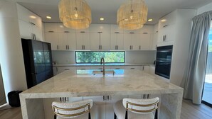 A modern kitchen with a large island, two pendant lights, and a view of greenery outside.