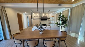 A modern dining room with a wooden table, four wicker chairs, a central plant, and a kitchen in the background.