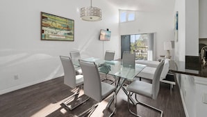 A modern dining area with a glass table and white chairs, a hanging light fixture, and artwork on the walls. - Cozy Apt w/ Pool, Hot Tub, Golf, Near Downtown! (Orlando)