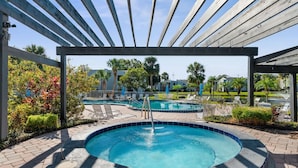 A circular hot tub with a tiled edge, surrounded by a wooden pergola, and a pool area with lounge chairs and palm trees in the background. - Cozy Apt w/ Pool, Hot Tub, Golf, Near Downtown! (Orlando)