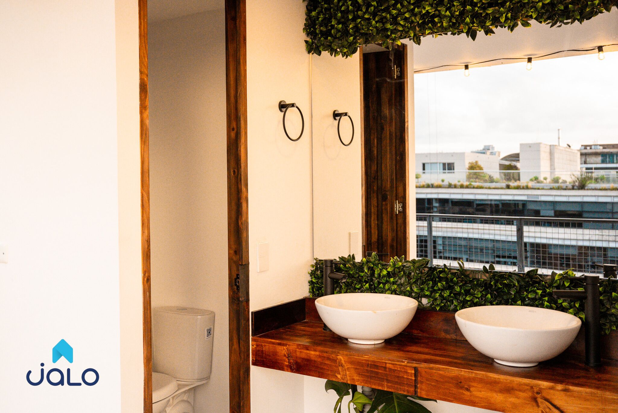 A modern bathroom with two large white sinks, a wooden countertop, a mirror, and a view of the cityscape through a window.